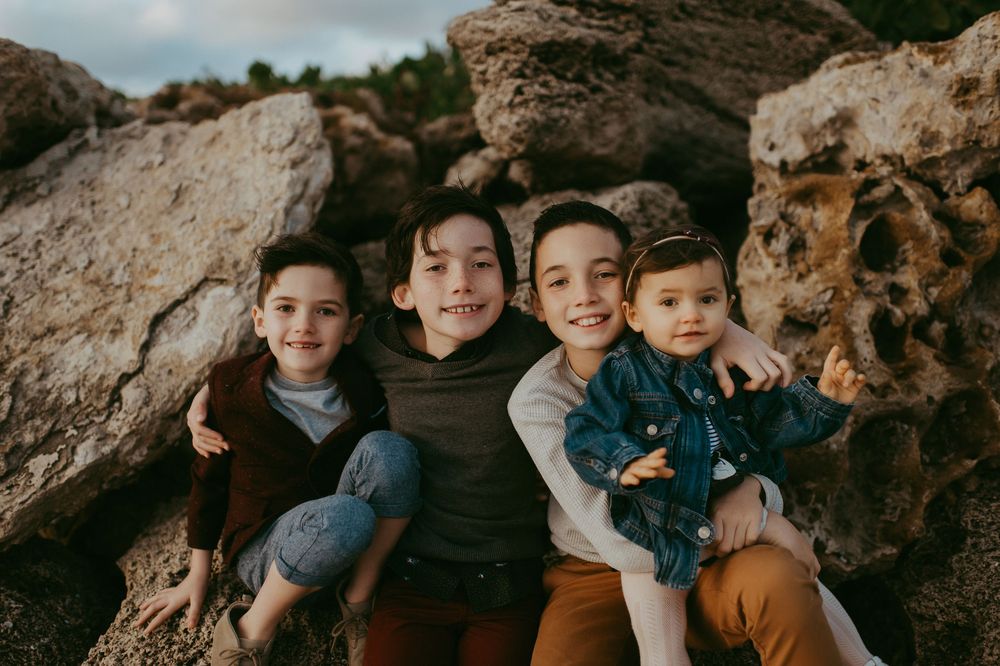 Four young siblings smile and hug together while sitting on large rocks outdoors.