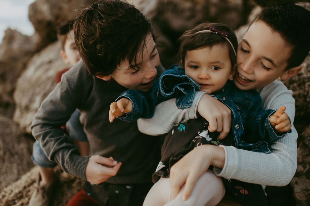 Two siblings lovingly hold and kiss a smiling baby girl outdoors near rocky terrain.