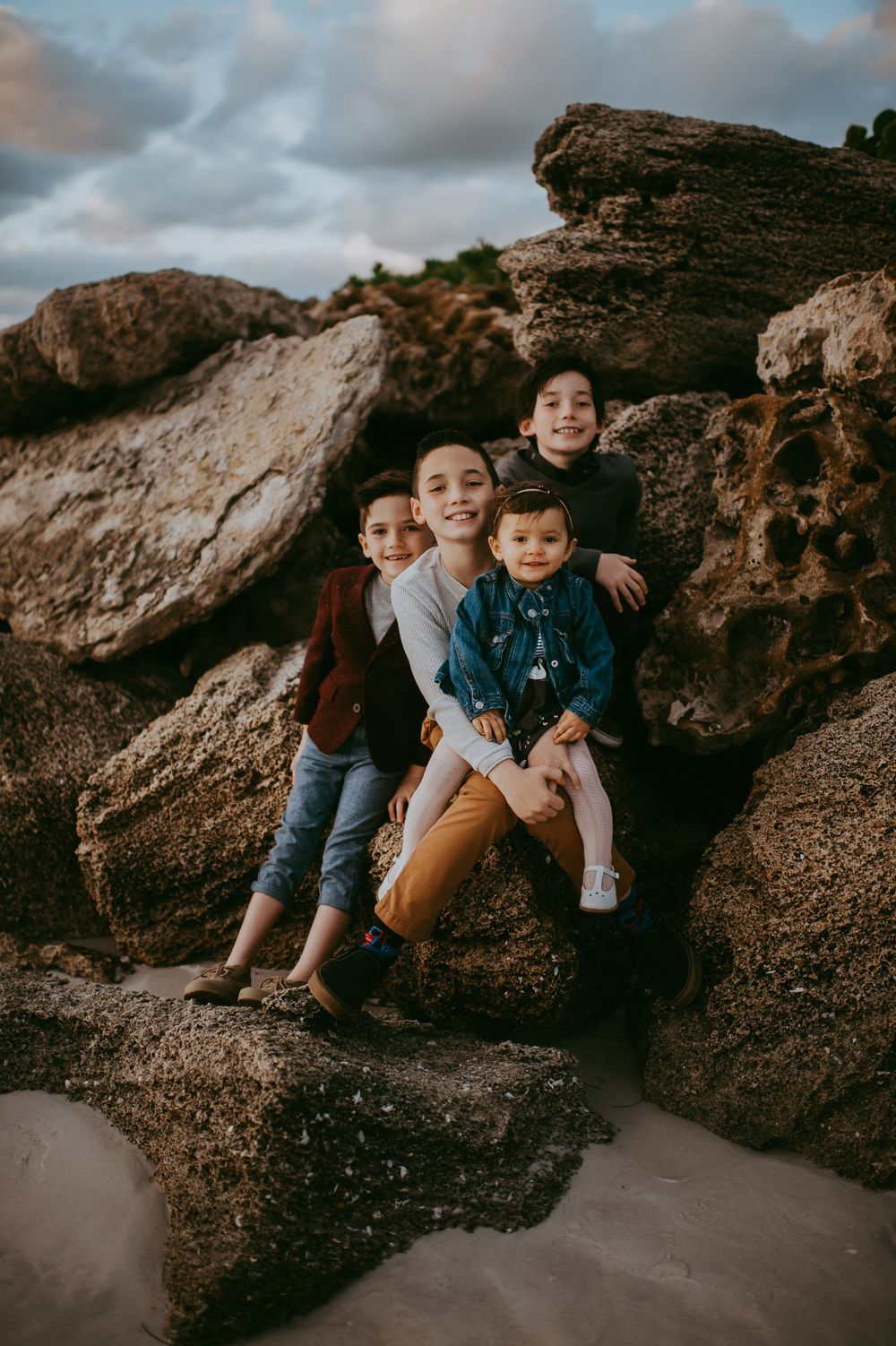Four children pose together on rocky beach boulders during a cloudy sunset family photo session.