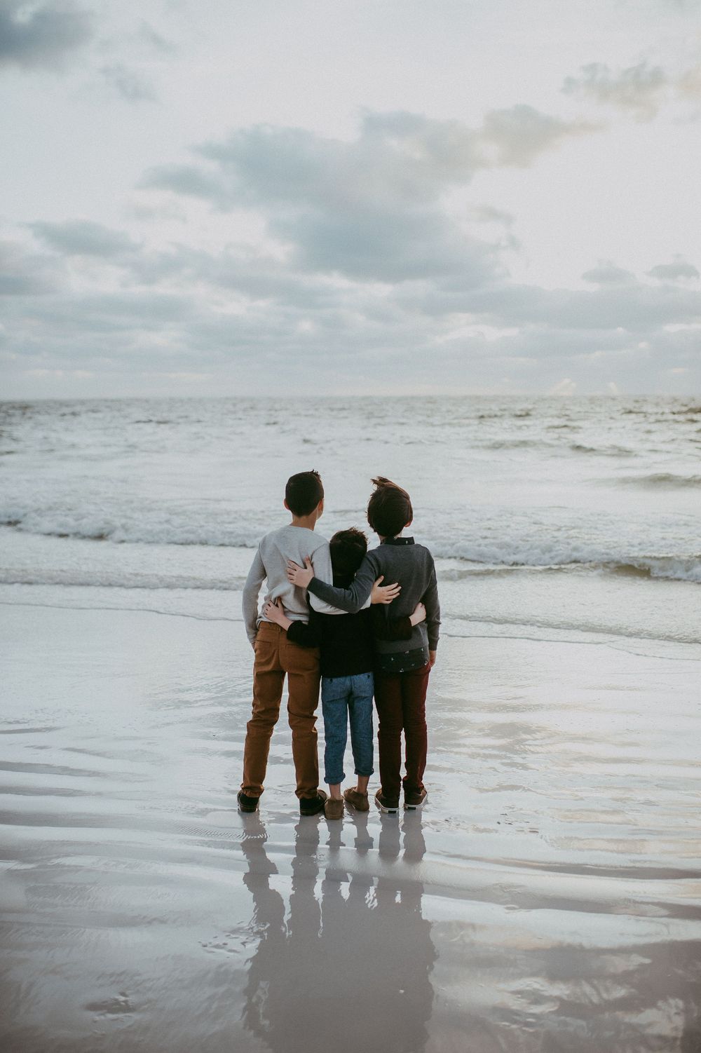 Three children stand embracing on a sandy beach, gazing at ocean waves under a cloudy sky.