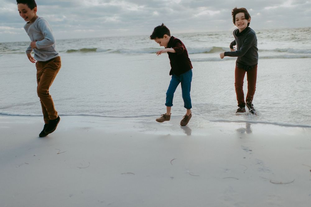 Three boys playing and running along a sandy beach with ocean waves in the background on a cloudy day.