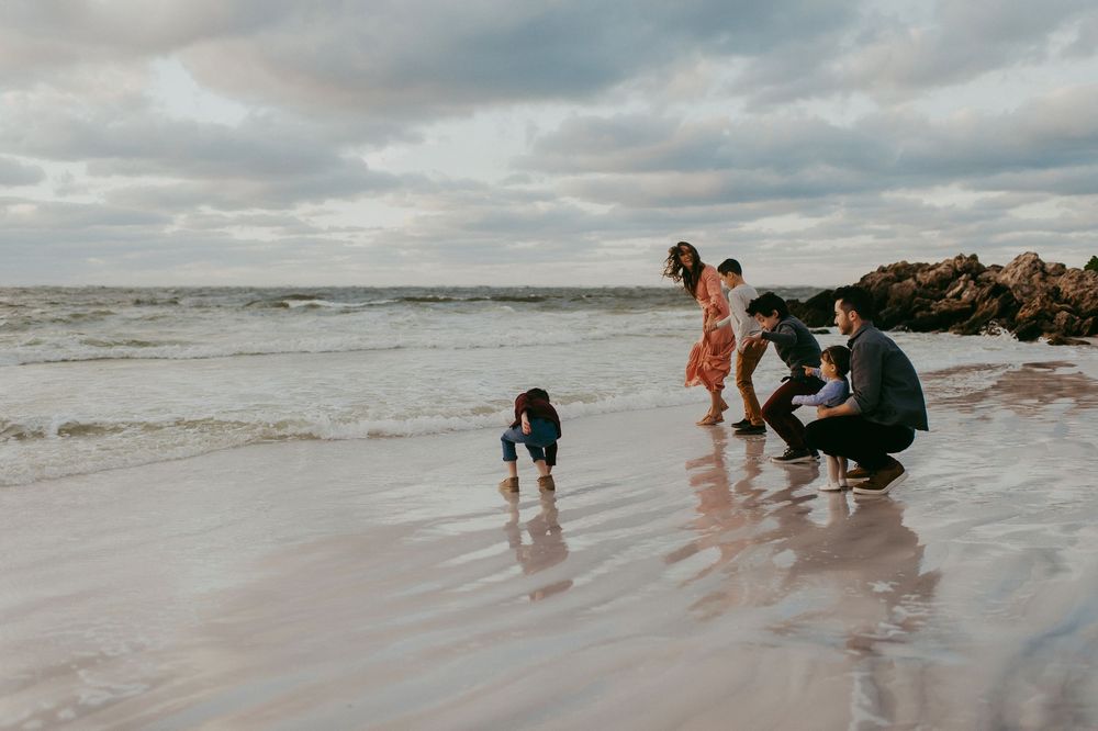 Family with young children exploring the shoreline on a rocky beach under a cloudy sky.