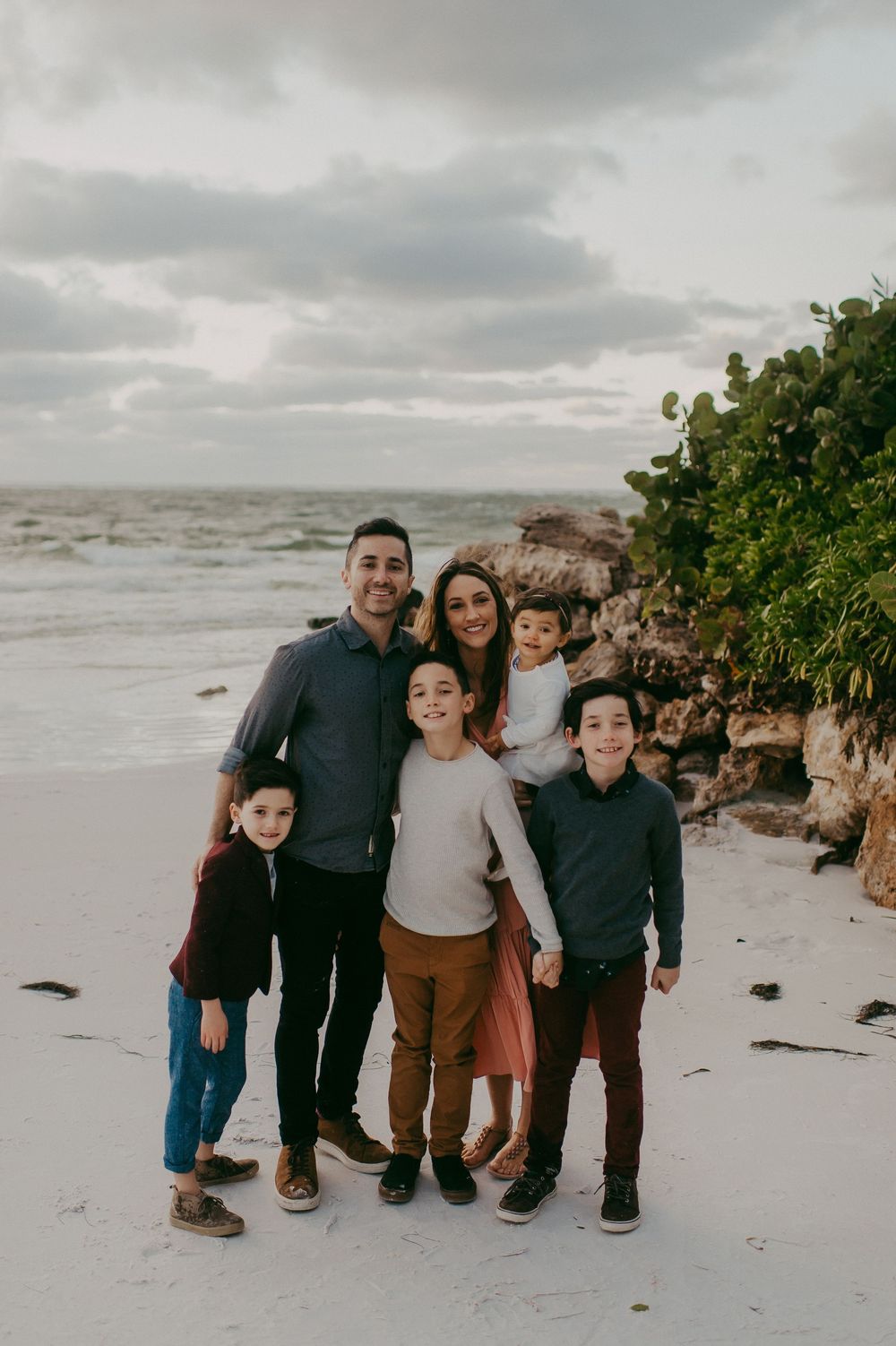 Happy family of six poses together on a sandy beach with ocean waves and rocky shore behind them.