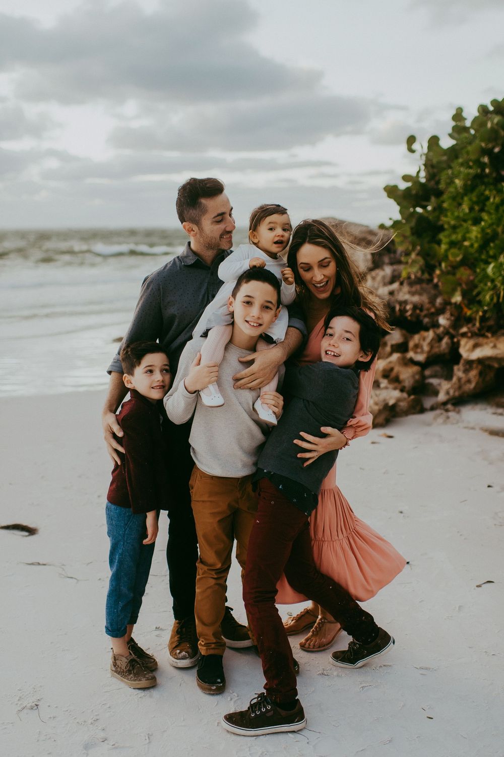 Happy family of six posing together on a sandy beach with ocean waves and green trees in the background.