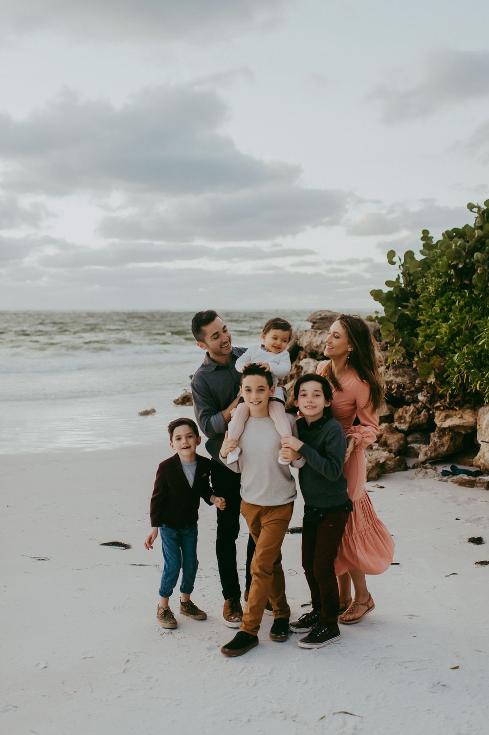 Family of six posing together on a sandy beach at dusk with cloudy skies and ocean waves in the background.