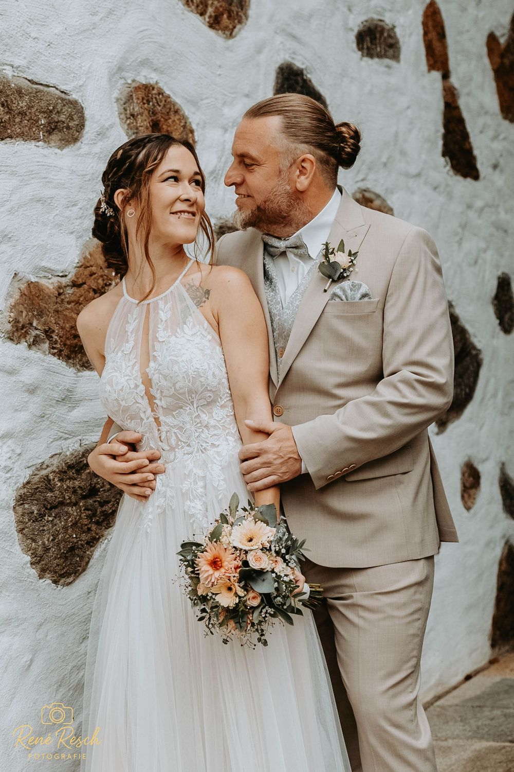 Bride in white gown holds floral bouquet beside groom in tan suit, posing against rustic stone wall.