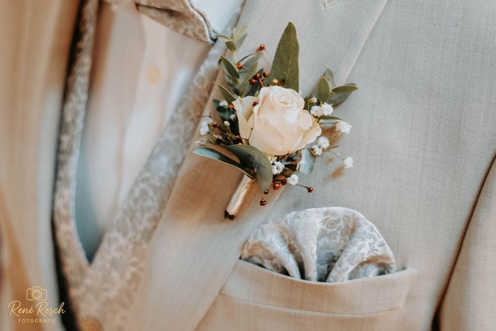 Close-up of a groom's boutonniere featuring a cream rose and greenery on a beige suit with a floral pocket square.