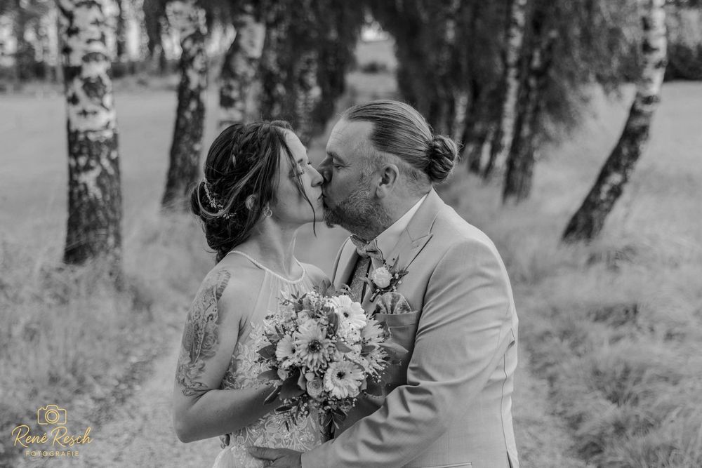 Black and white photo of a bride and groom sharing a kiss outdoors among birch trees, bride holding a floral bouquet.