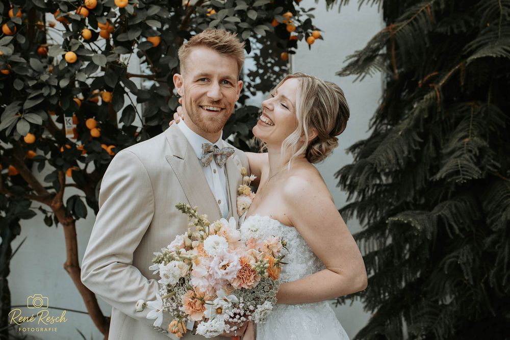 A smiling bride and groom pose together outdoors, she holds a peach floral bouquet, he wears a beige suit with bow tie.