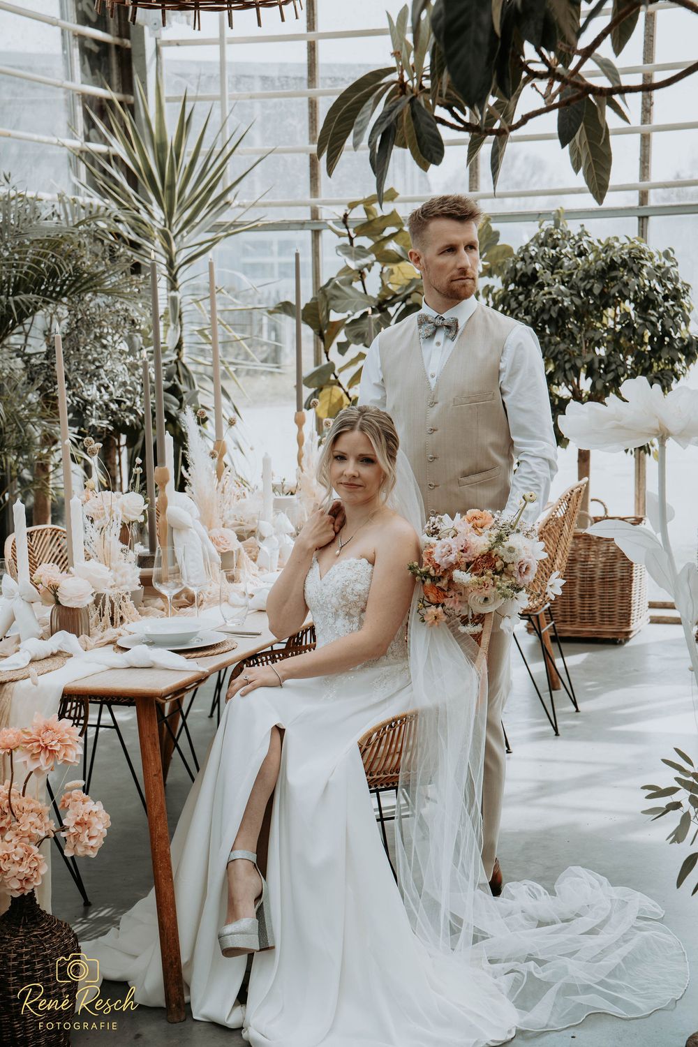 Bride in white gown seated at boho greenhouse wedding table with groom standing behind her holding bouquet.