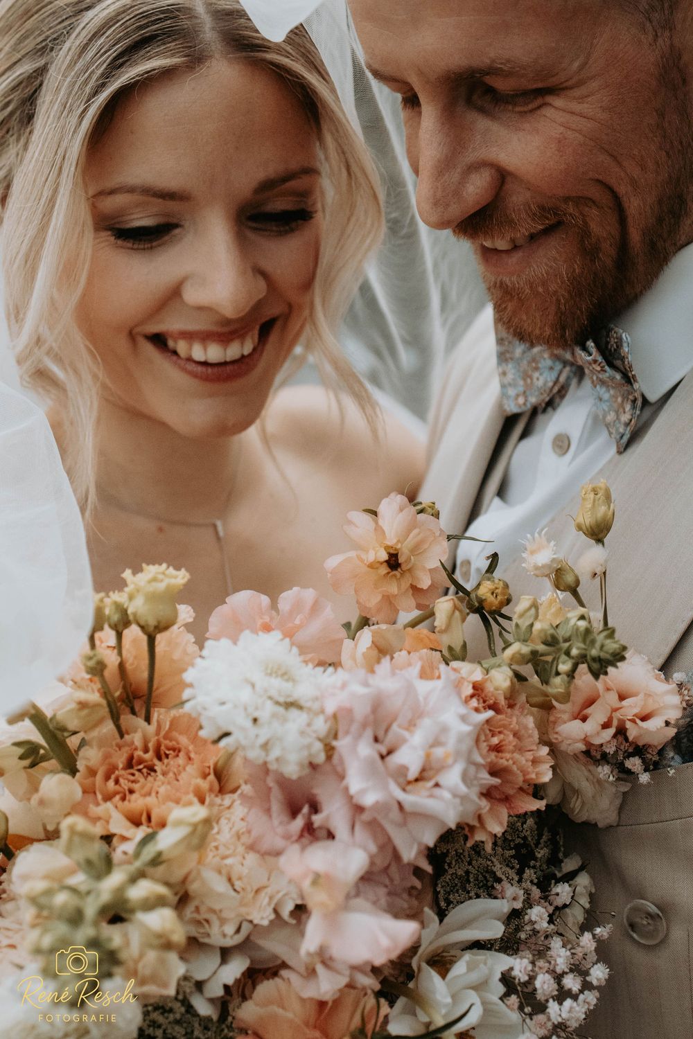 Smiling bride holding a pastel pink and peach wedding bouquet with carnations beside a bearded groom in a suit.
