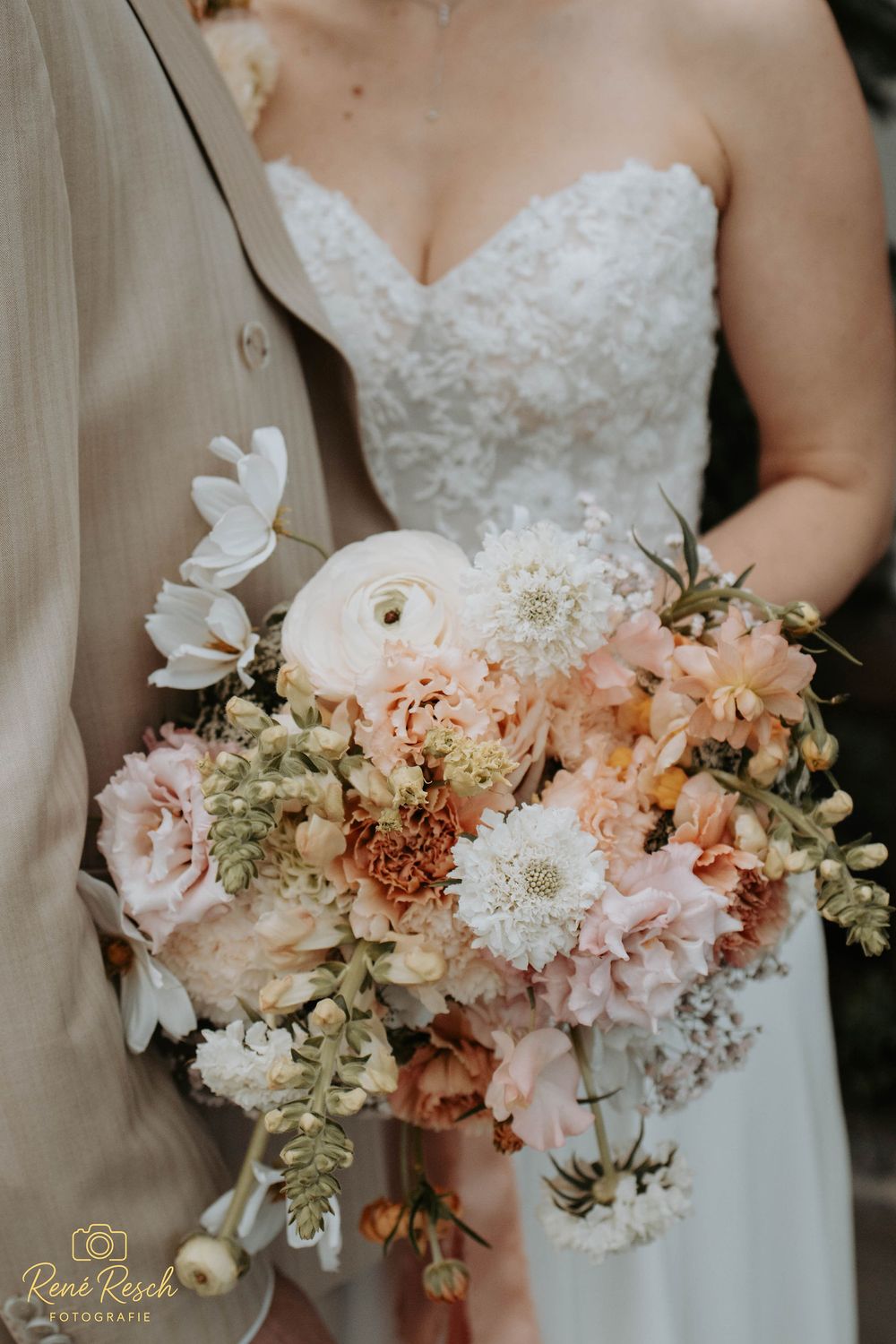 Bride holding a lush wedding bouquet with blush, peach, and white flowers including ranunculus and dahlias, standing beside the groom.