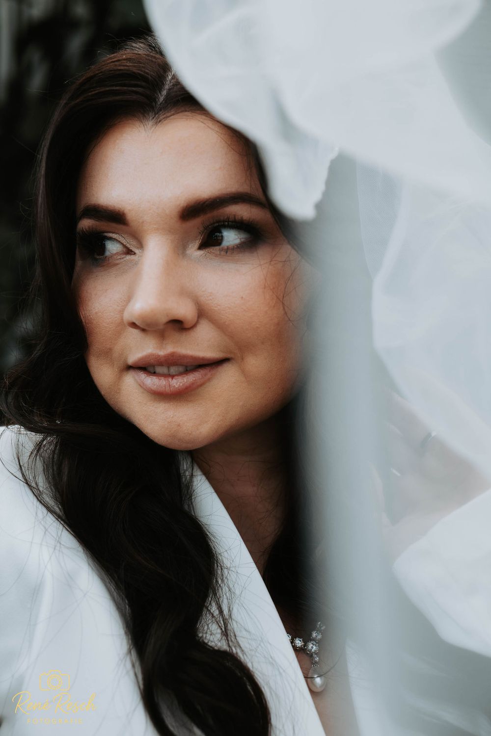 A bride with dark hair and natural makeup gazes upward, framed by flowing white veil fabric.