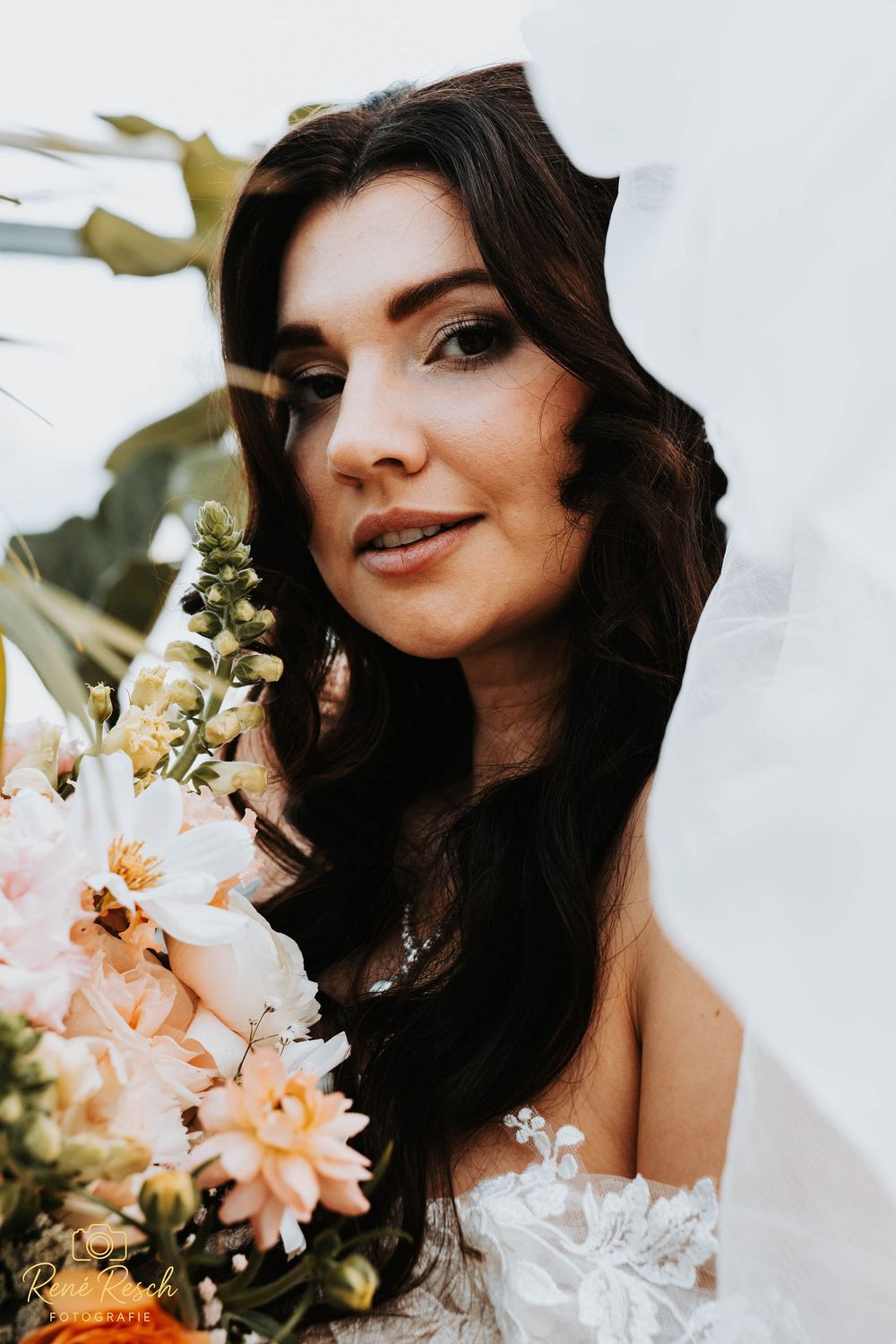 Bride with dark hair holding a peach and white floral bouquet, wearing a lace wedding dress, soft natural light.