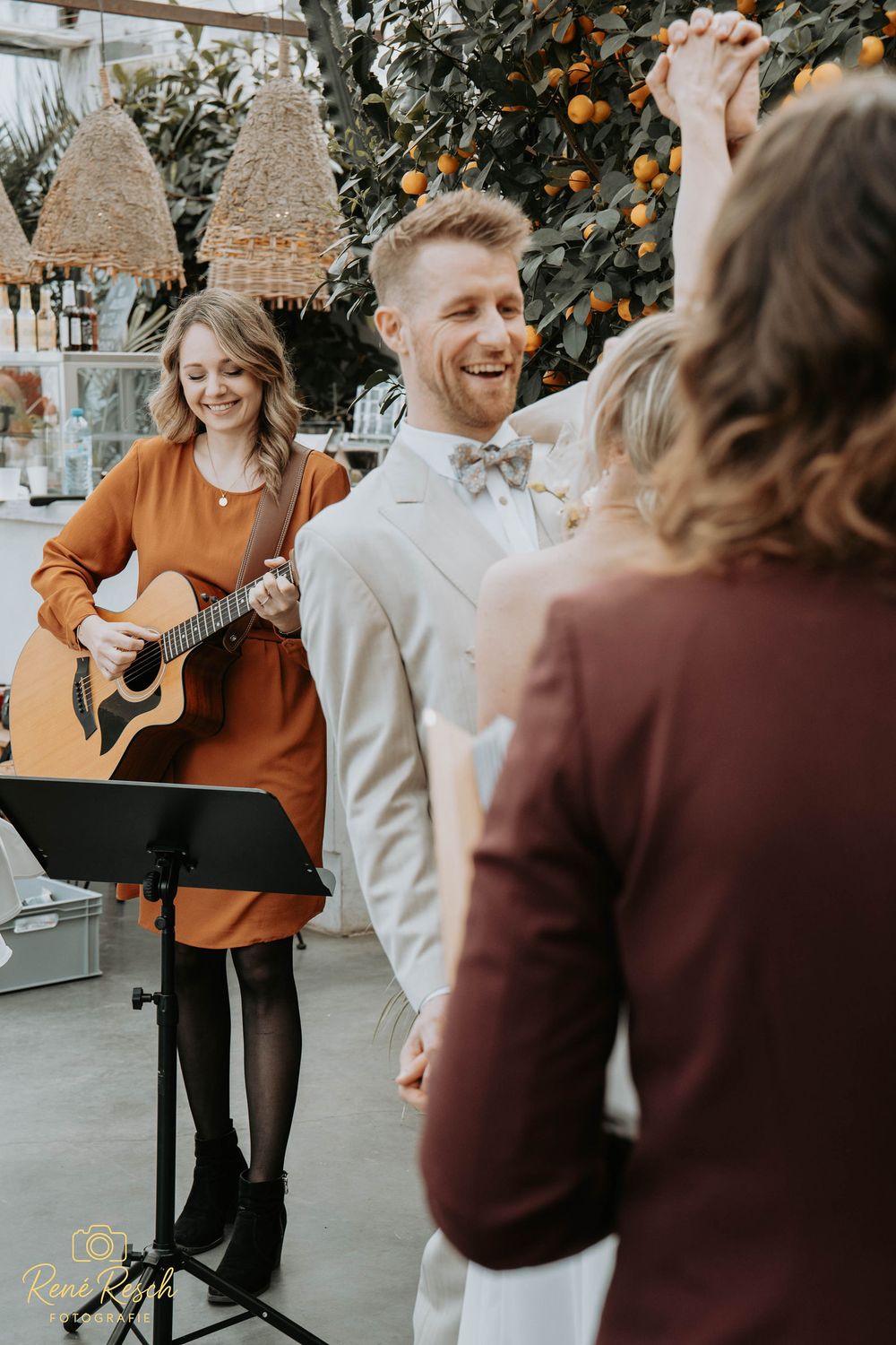 A woman in an orange dress plays acoustic guitar at a wedding ceremony while a groom in a gray suit laughs joyfully.