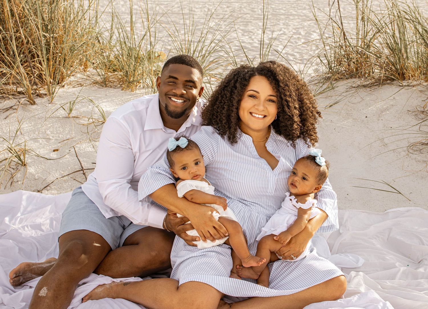 Happy family of four posing on a sandy beach, parents holding twin babies in white outfits near sand dunes.