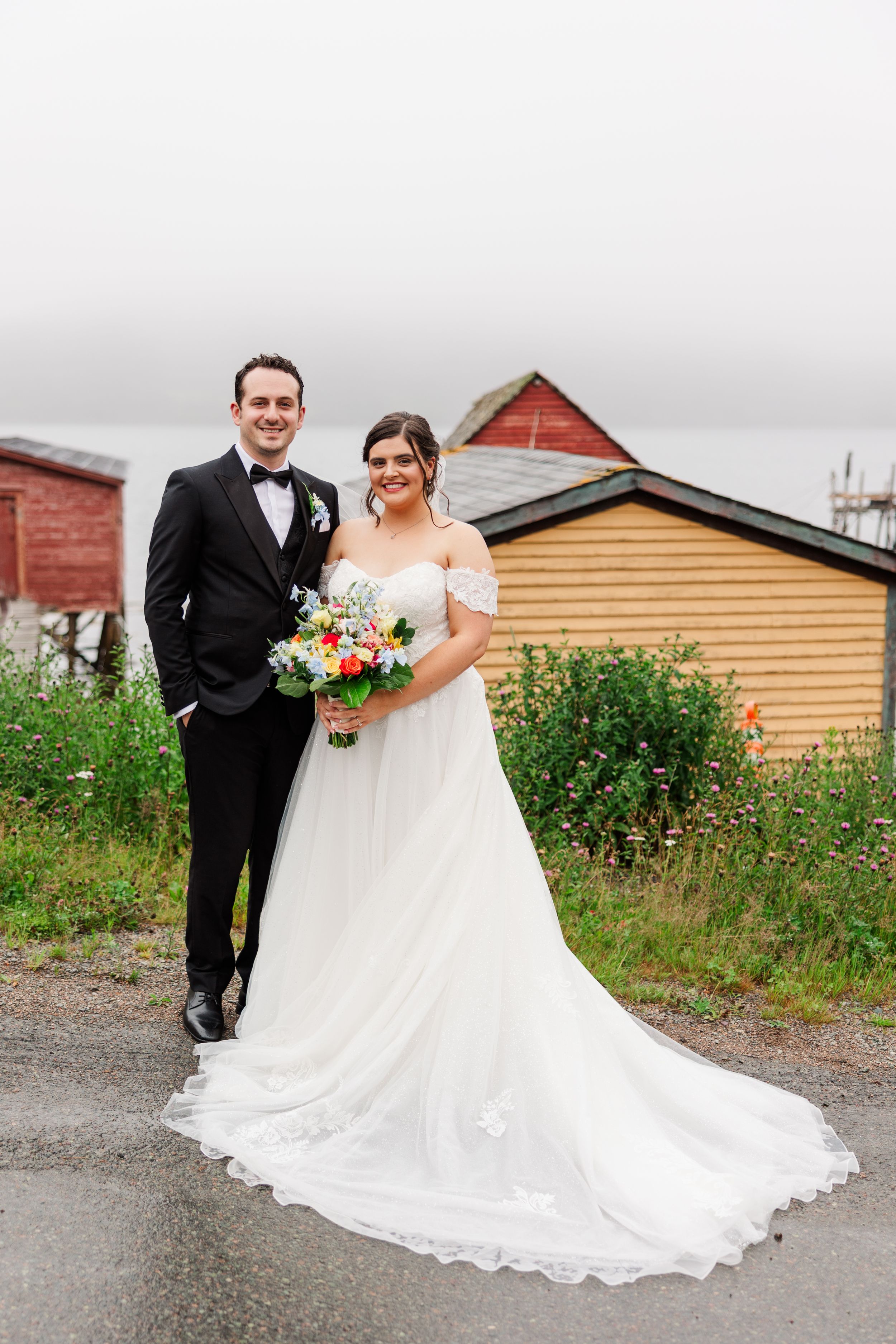 Bride in white off-shoulder gown holding colorful bouquet stands with groom in black tuxedo outside rustic buildings.
