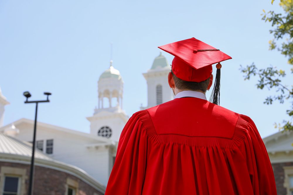 Graduate in red cap and gown stands facing a white historic building on a sunny graduation day.