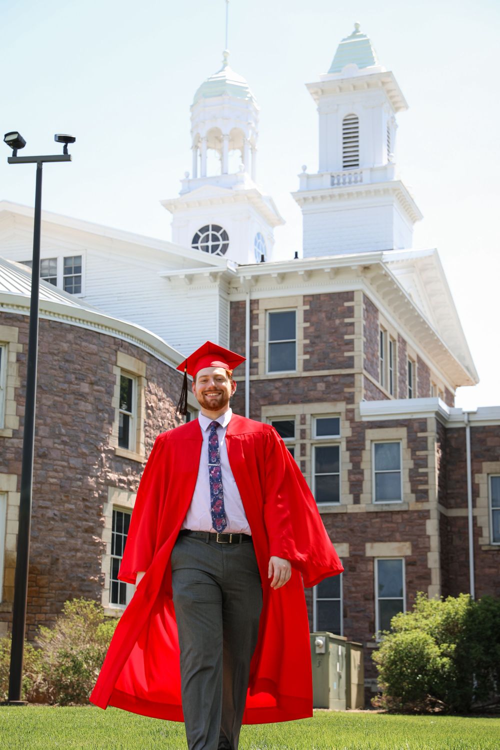 Smiling male graduate in red cap and gown walks across campus lawn in front of historic brick building with white towers.