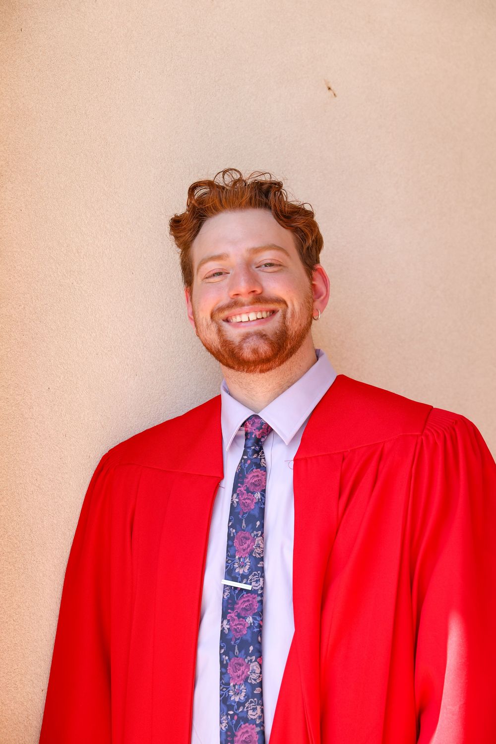 Smiling young man with red beard wearing red graduation gown and floral tie against beige wall.