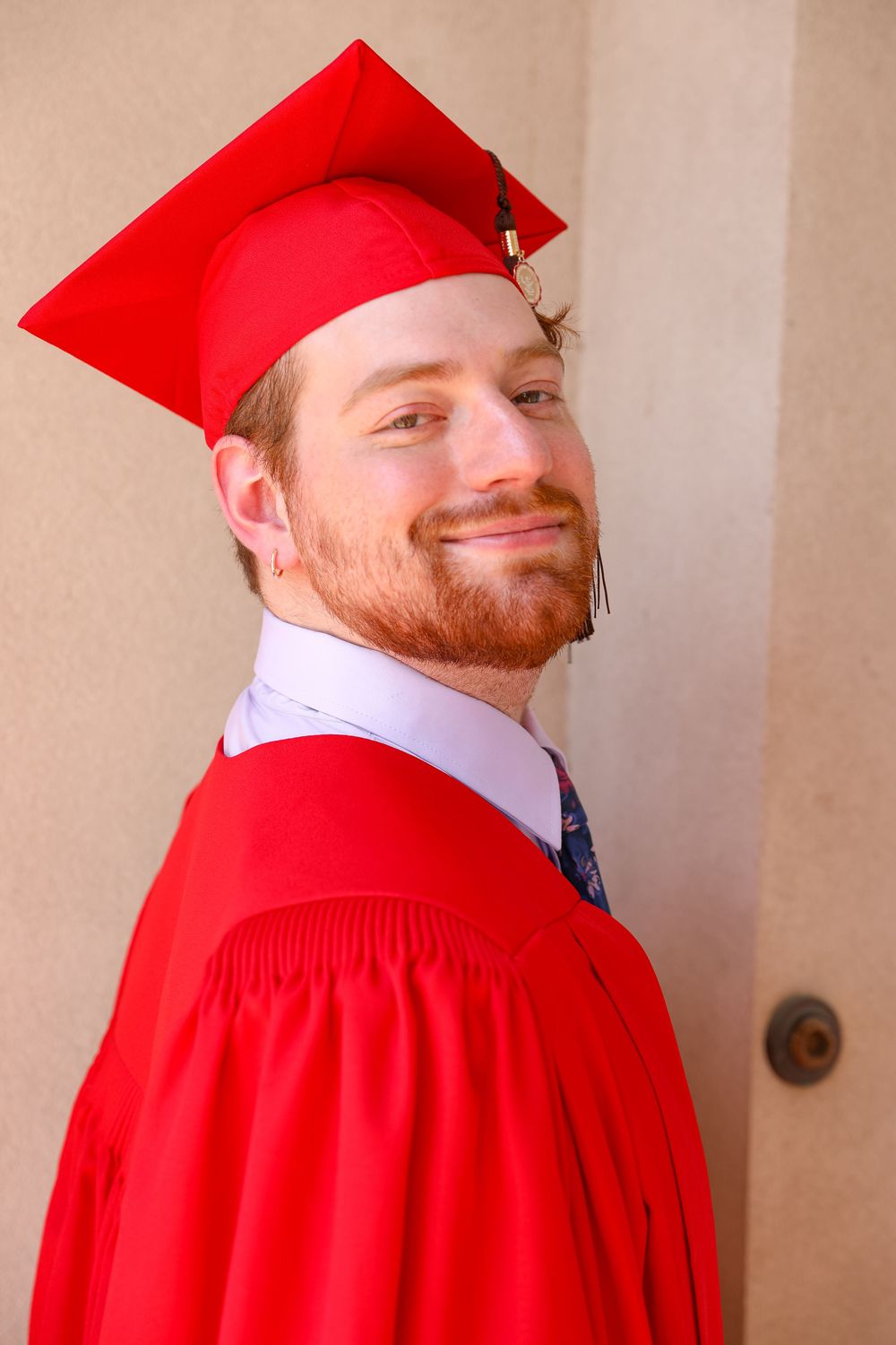 Young man with red beard smiles in red graduation cap and gown posed against a neutral wall outdoors.