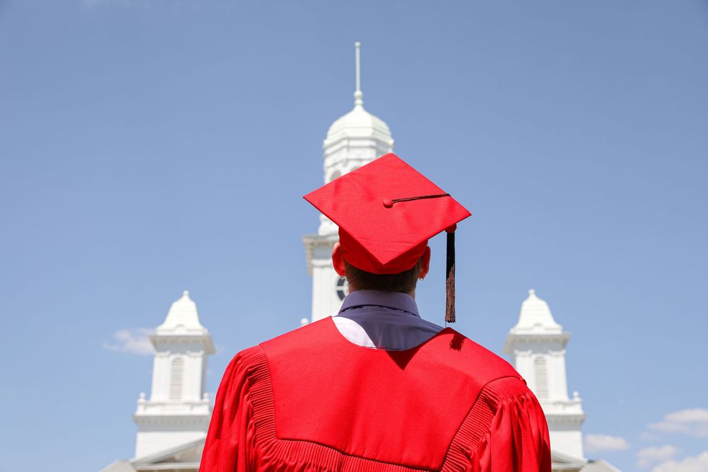 Graduate in red cap and gown stands before white building with spires under blue sky.