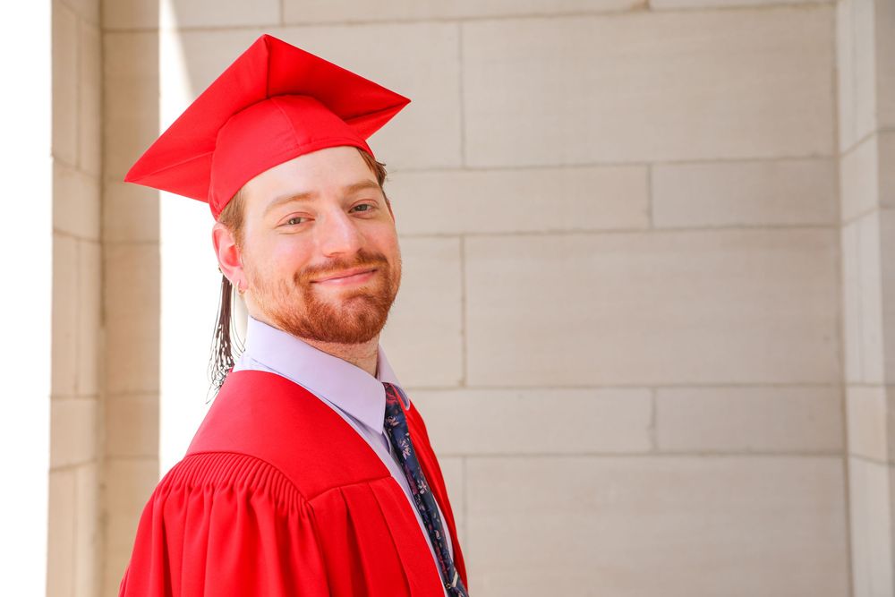 Smiling male graduate in red cap and gown posing outdoors against a stone wall background.