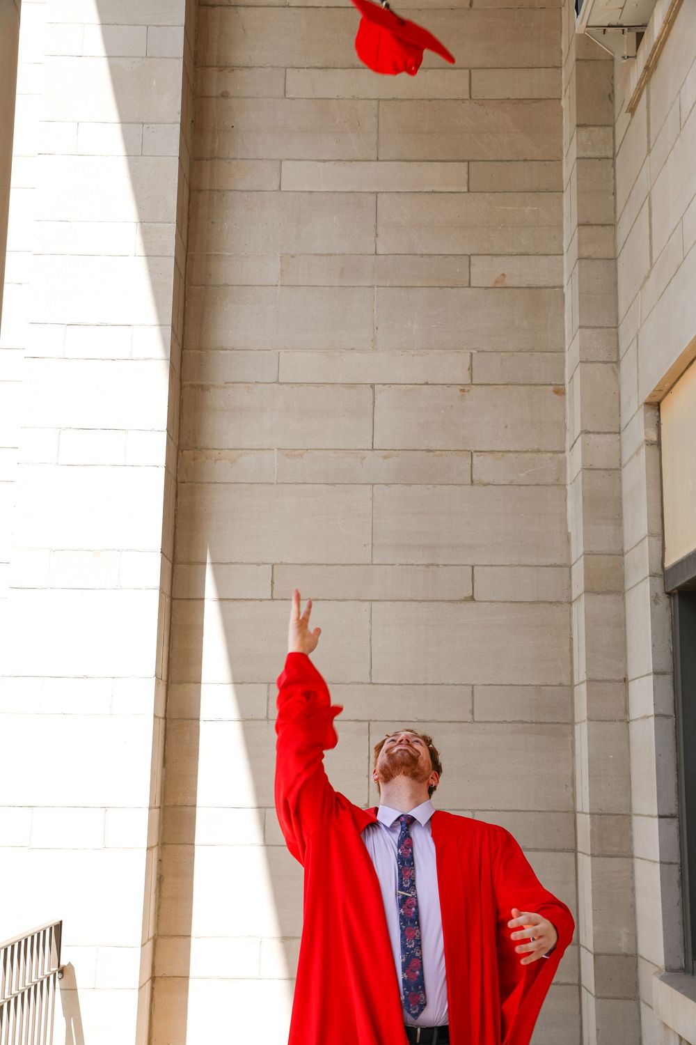 Graduate in red gown tossing mortarboard cap in the air outdoors near stone building columns.