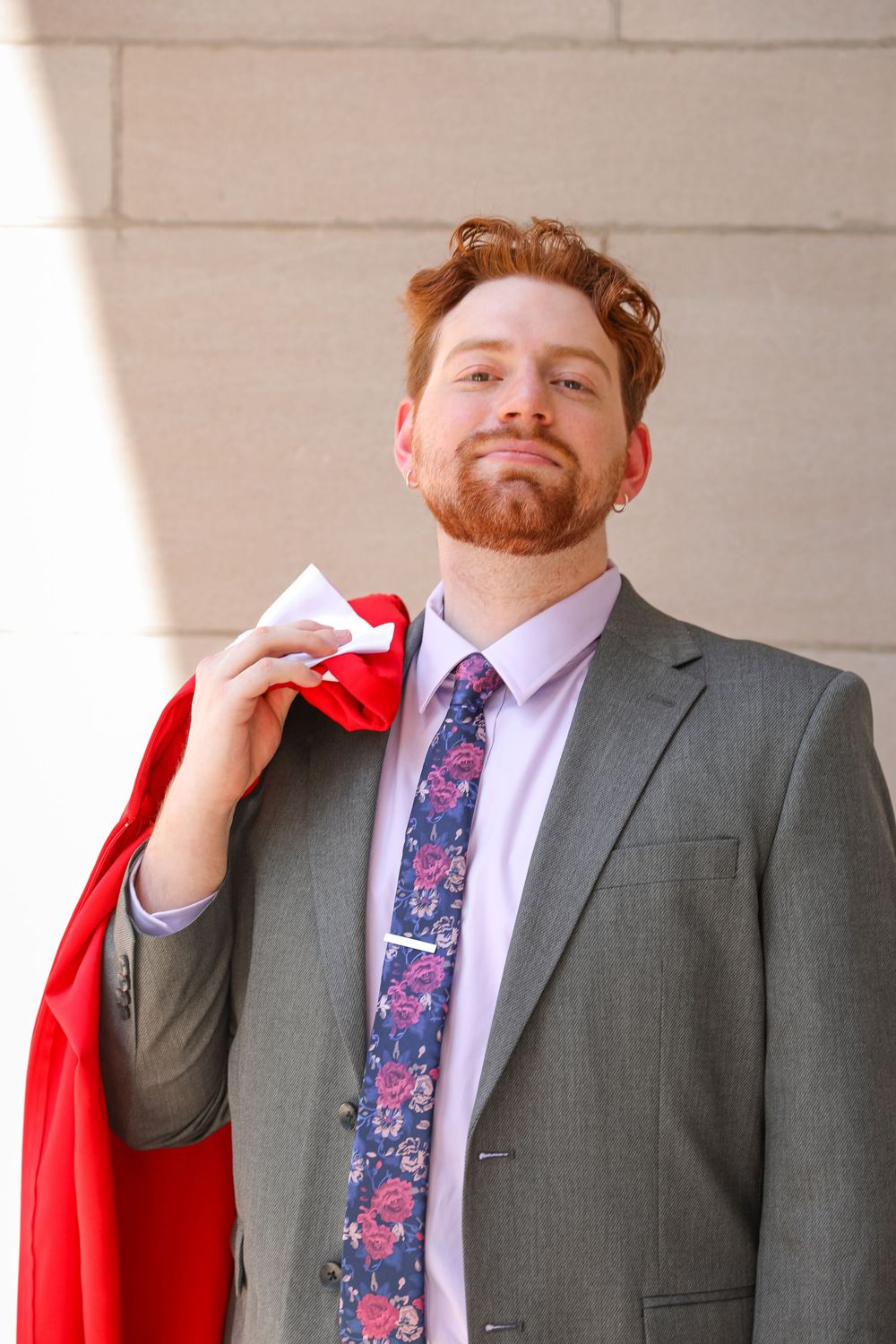 Confident redhead businessman in gray suit and floral tie holding red cape over shoulder against light wall.