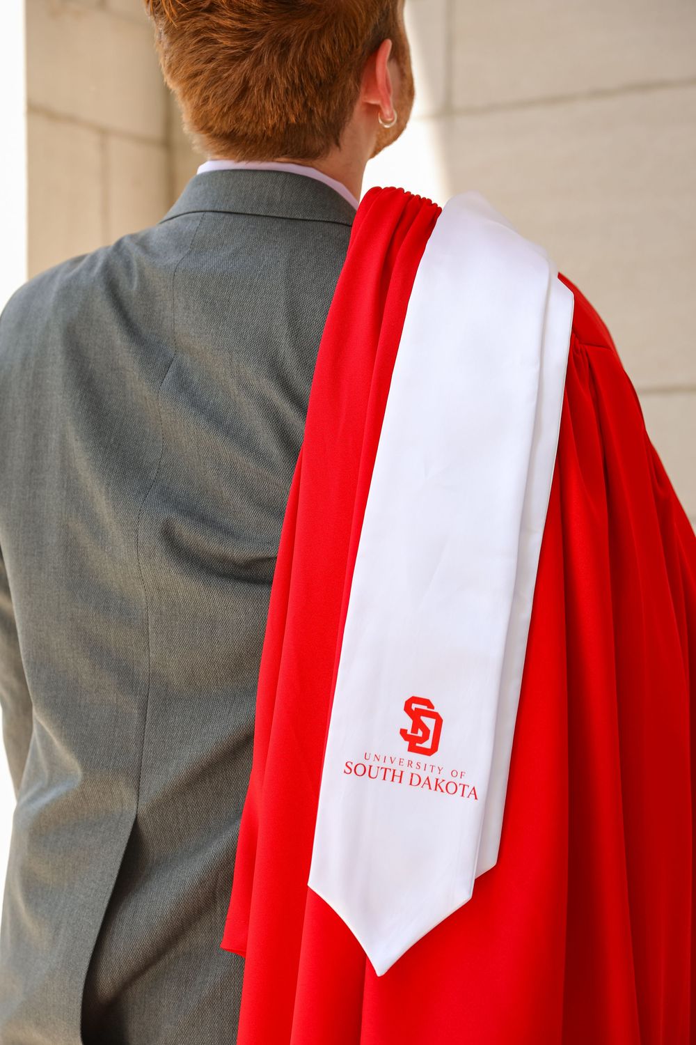 University of South Dakota graduate in red gown and white stole, viewed from behind, wearing a gray suit.