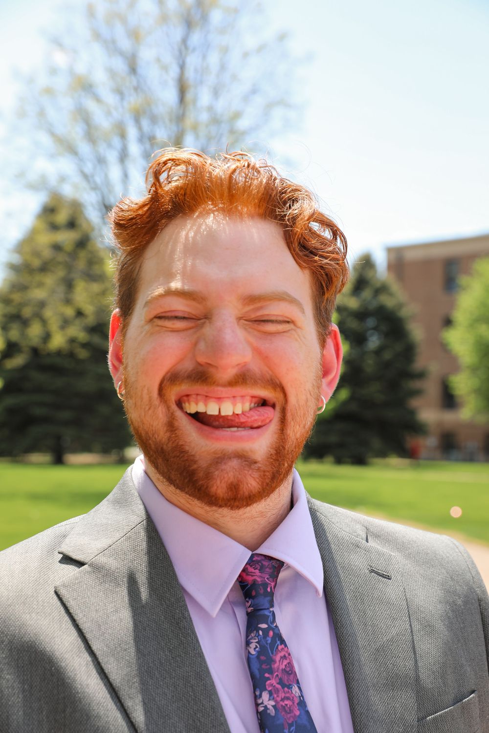 Laughing redhead man with beard wearing gray suit and floral tie outdoors on sunny day.