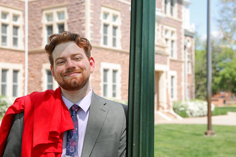 Young man with beard in gray suit holding red graduation gown, smiling outdoors near brick university building.