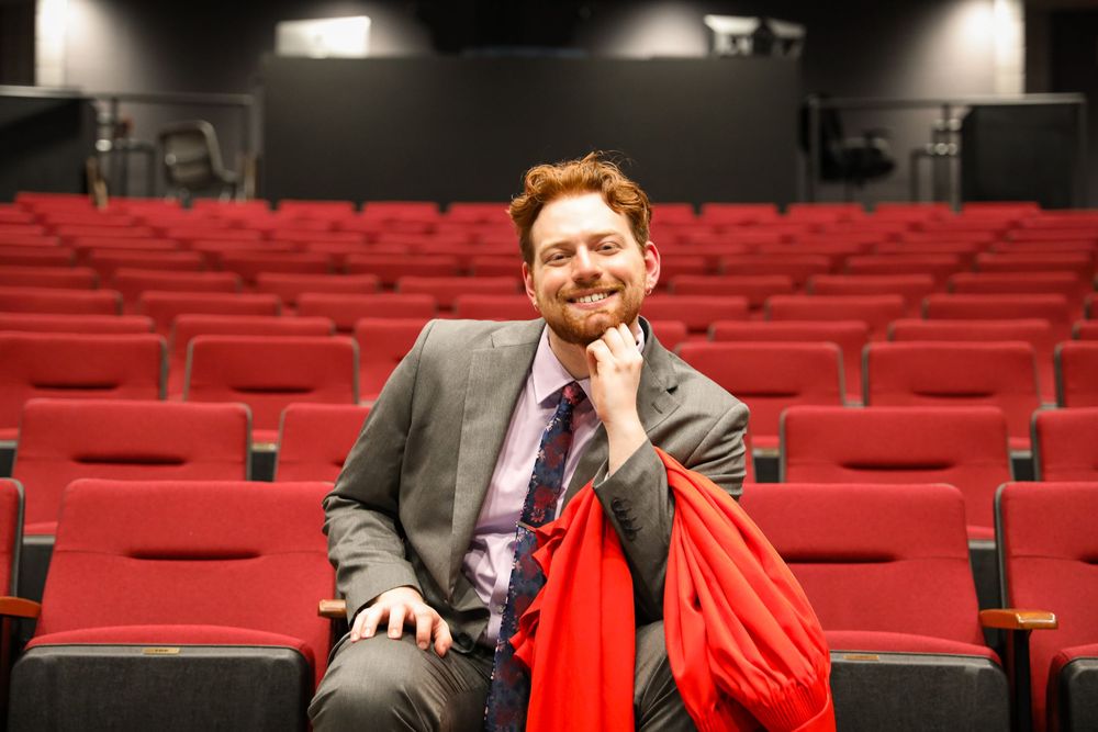 Smiling man in gray suit with red scarf sits relaxed in red theater auditorium seating.
