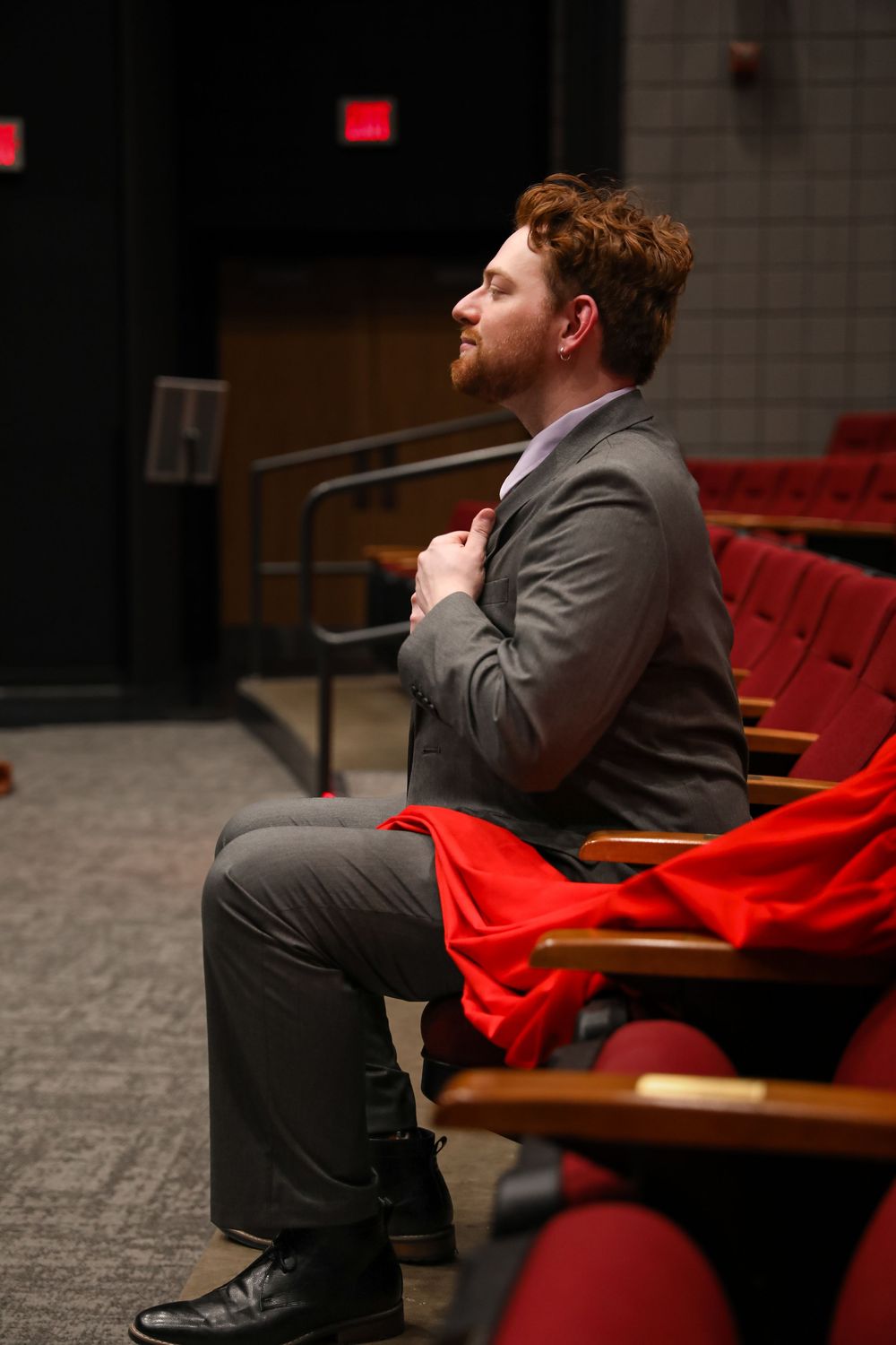 Bearded redhead man in gray suit sits in red theater seats, holding chest, with red cape draped over chair.