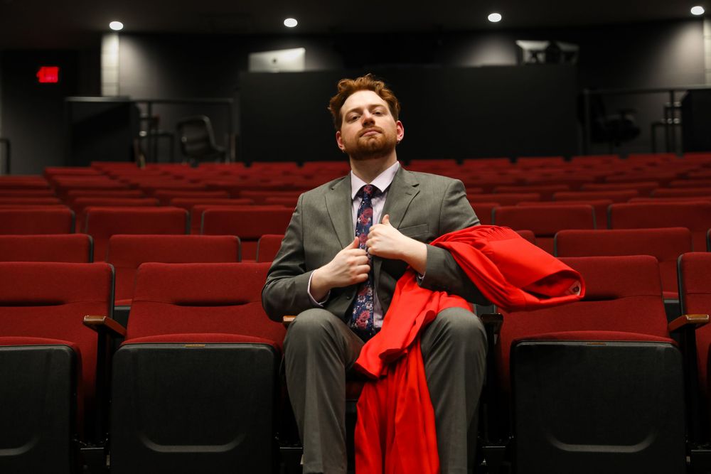 Man in gray suit holding red jacket sits alone in empty red theater seats.