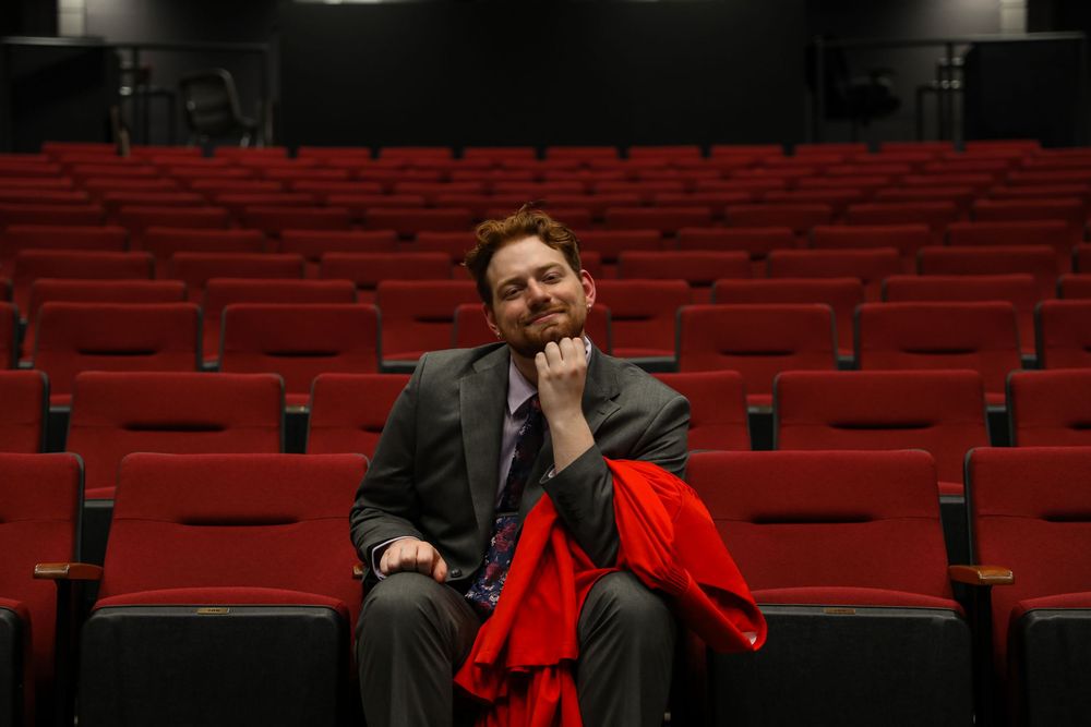 Man in gray suit sitting alone in red theater seats, resting chin on hand with red jacket draped over arm.