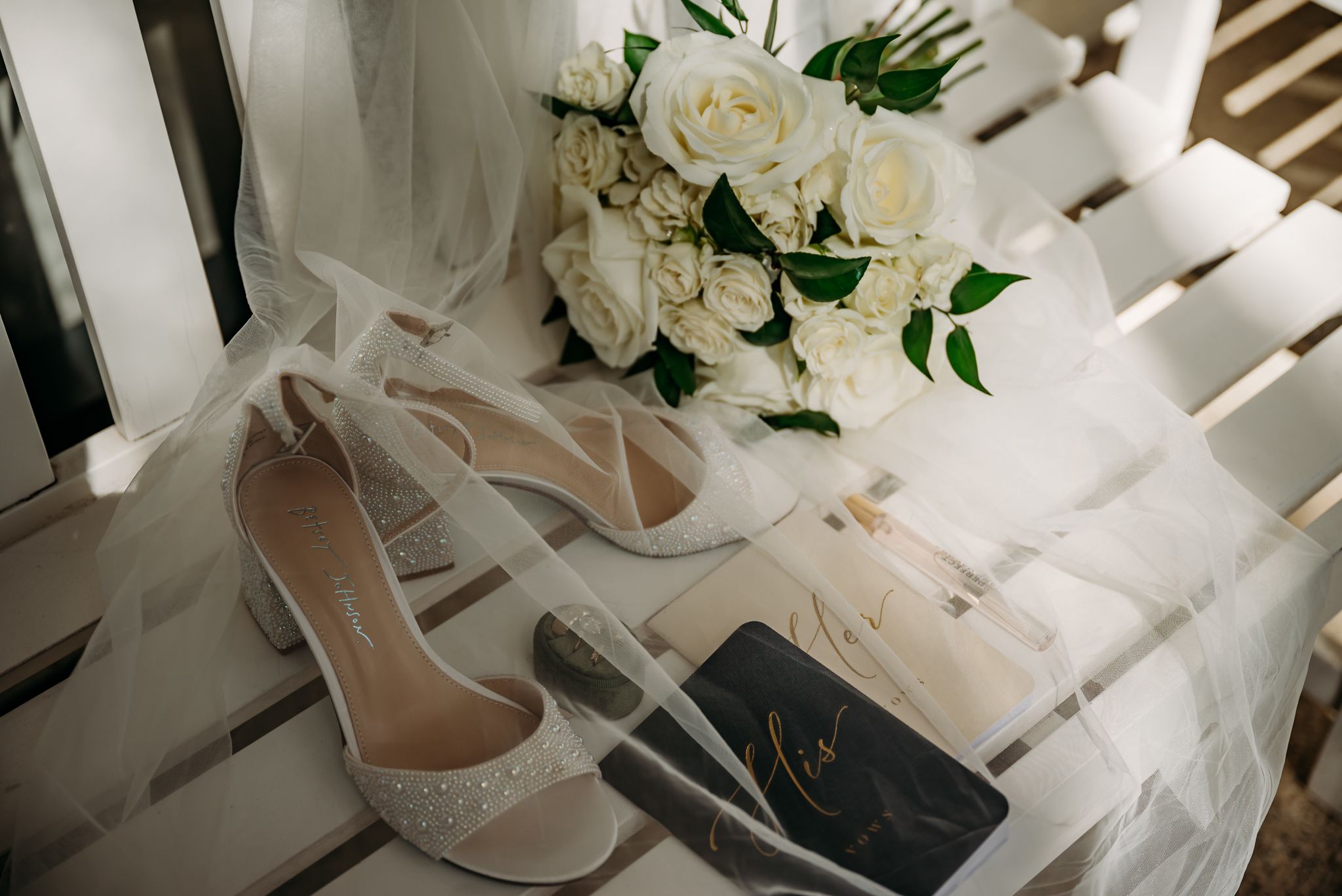 Elegant bridal flat lay featuring white heels, a lush white rose bouquet, sheer veil, and a dark wedding clutch on white chairs.