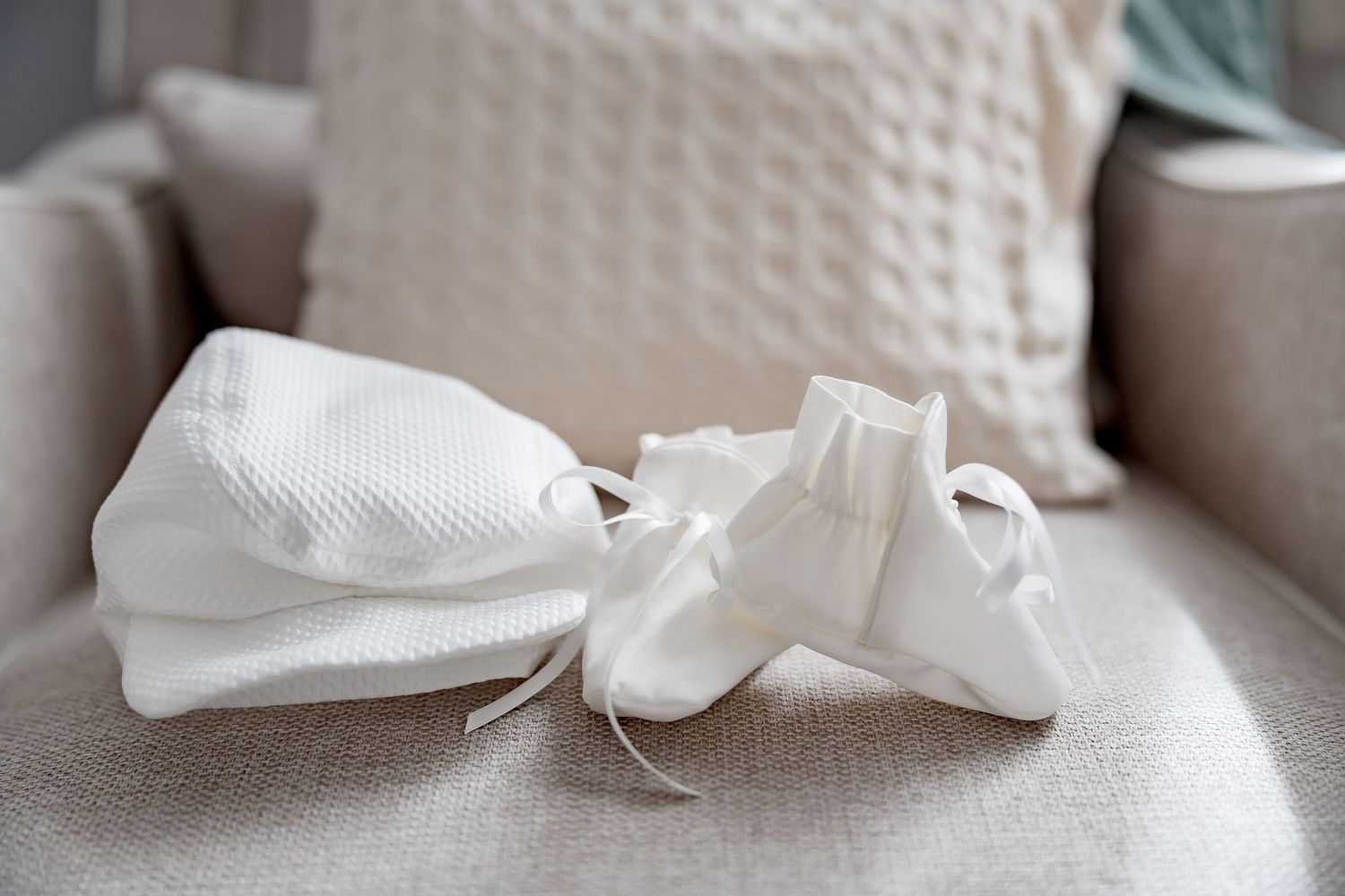 White baby baptism outfit with booties and hat laid on a beige chair for a christening ceremony.