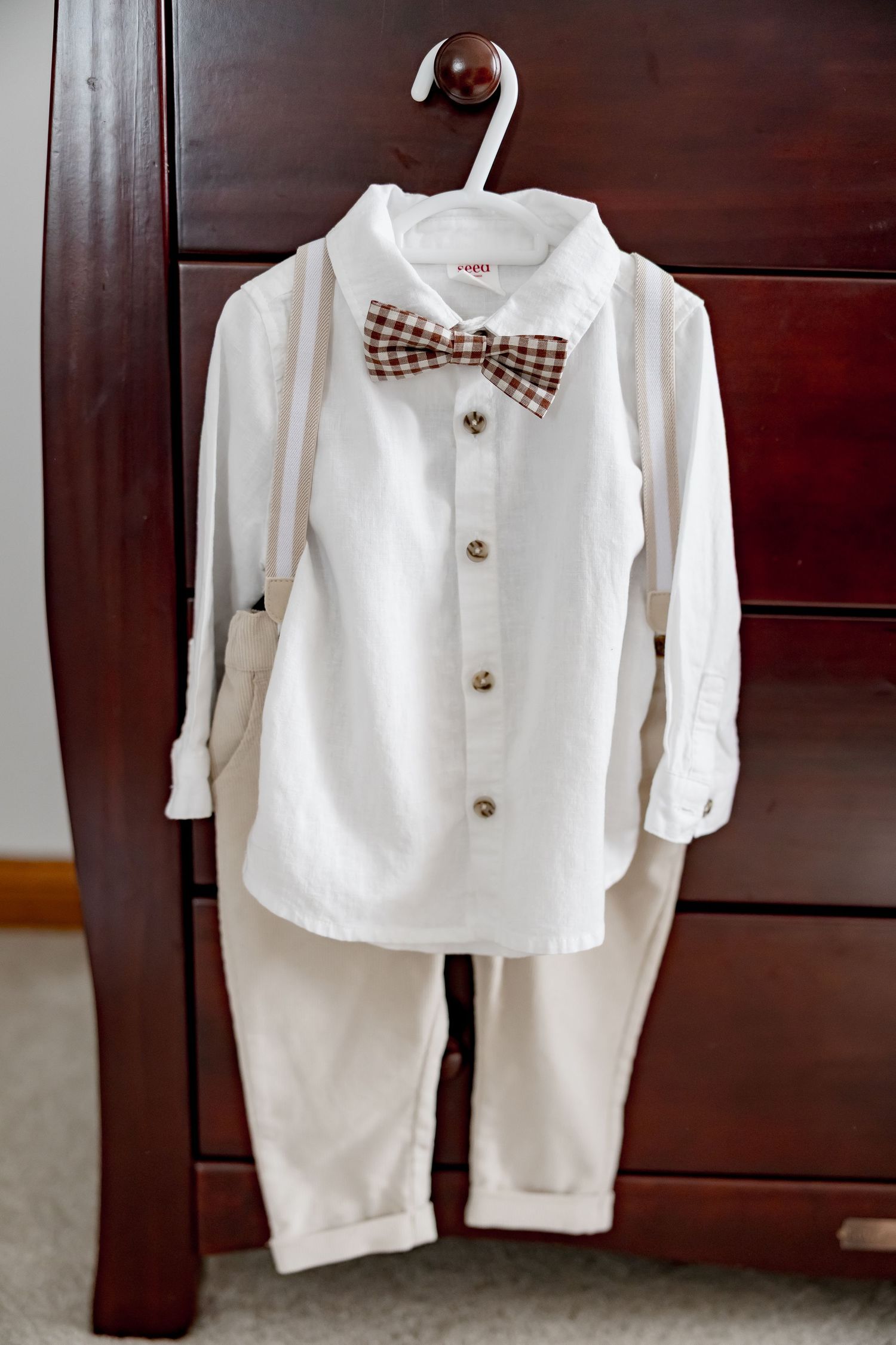 White baby baptism outfit with bow tie and suspenders hanging on wooden dresser.