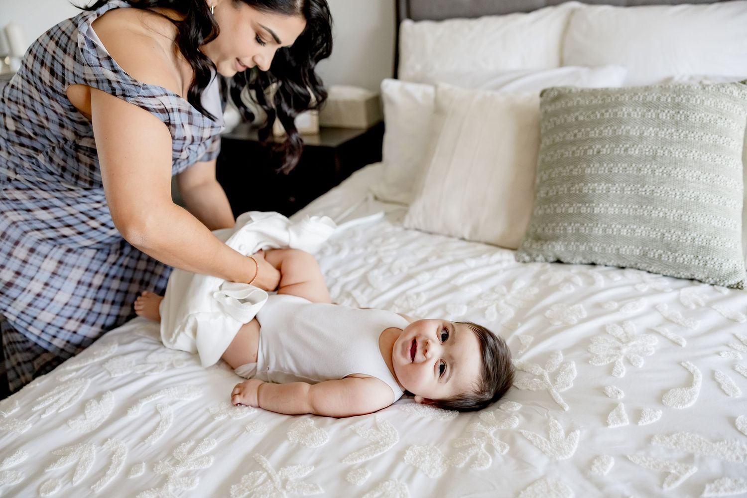 Mother changing baby diaper on white bed while baby smiles happily.