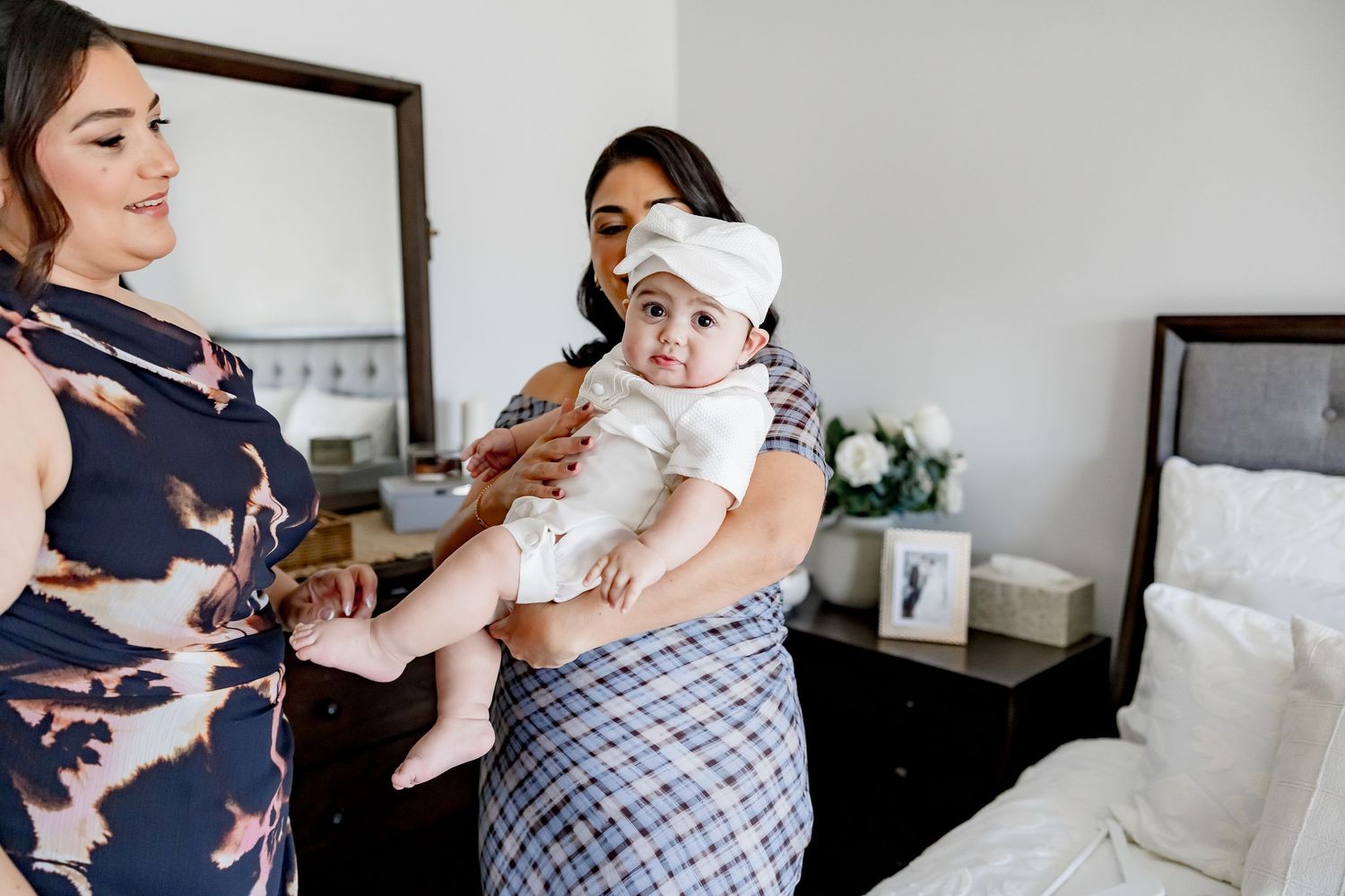 Two women in elegant dresses holding a baby in white baptism outfit in a bedroom.