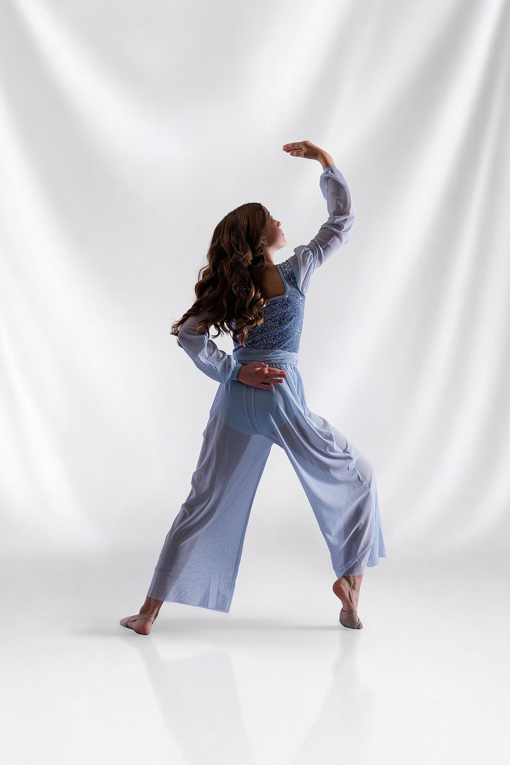 Young female dancer in black studded dress poses gracefully in dance studio with white draped backdrop.