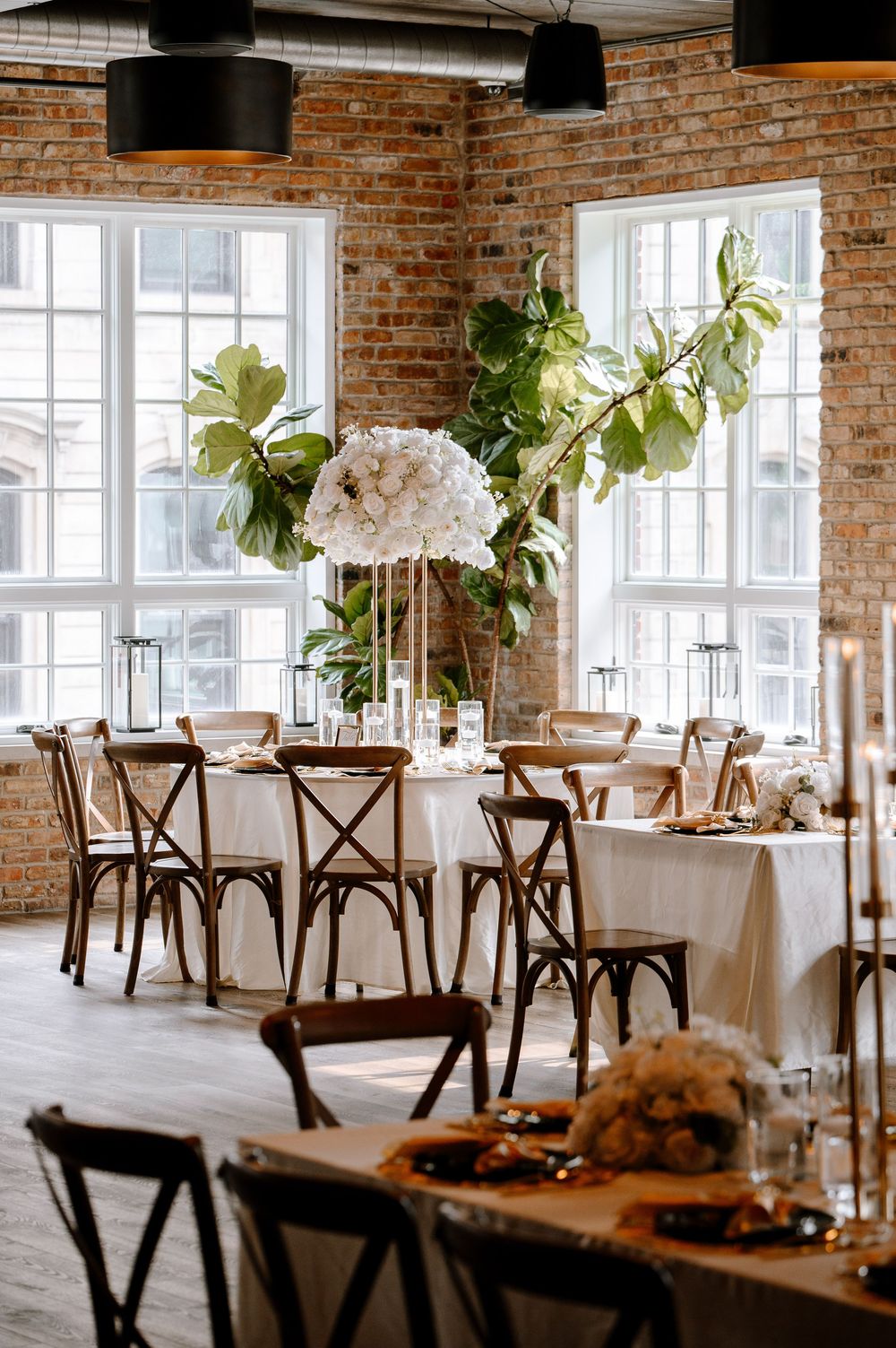 Elegant wedding reception with white floral centerpieces, cross-back chairs, and exposed brick walls in an industrial venue.