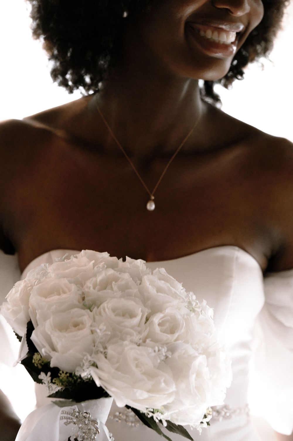 Smiling Black bride in strapless white gown holding a bouquet of white roses with a pearl necklace.