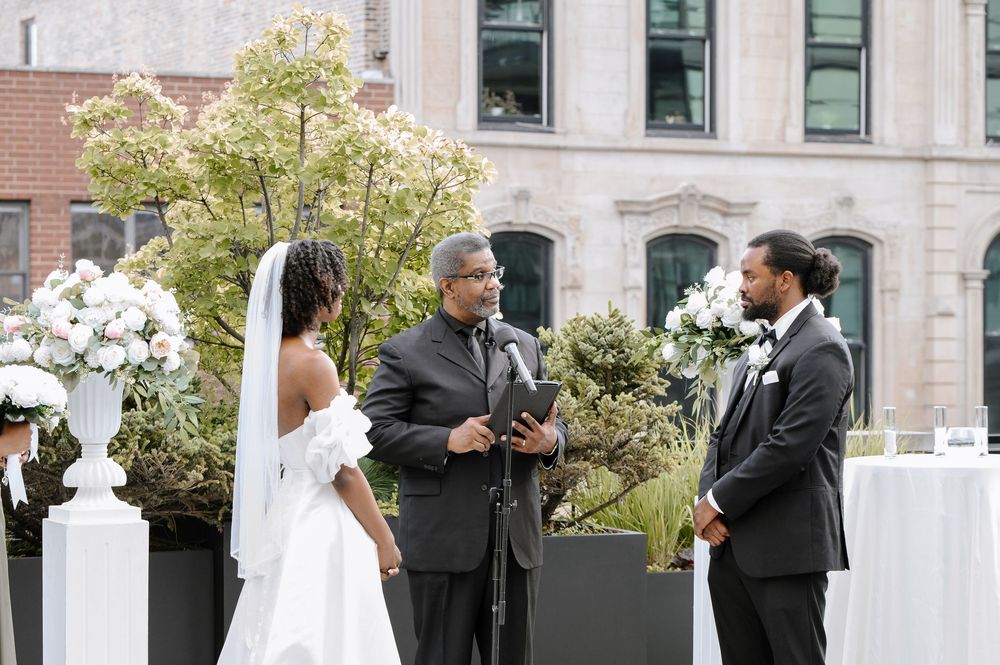 Outdoor wedding ceremony with bride, groom, and officiant at an elegant urban venue with white floral arrangements.