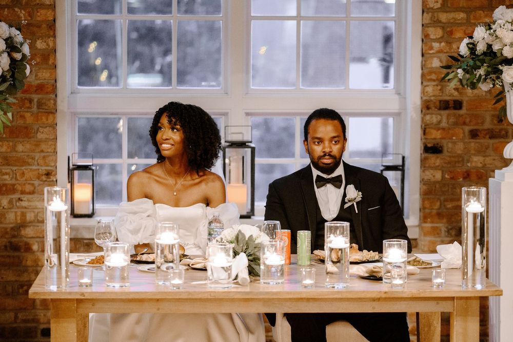 Elegant Black couple sits at sweetheart table with candles and floral decor at their wedding reception.