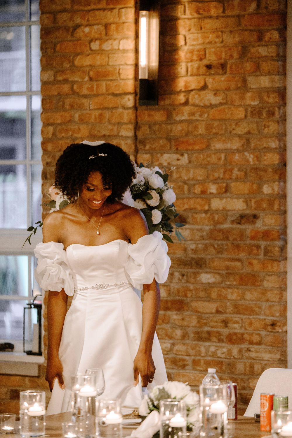 Radiant Black bride in off-shoulder puff sleeve wedding gown smiling near candlelit reception table.