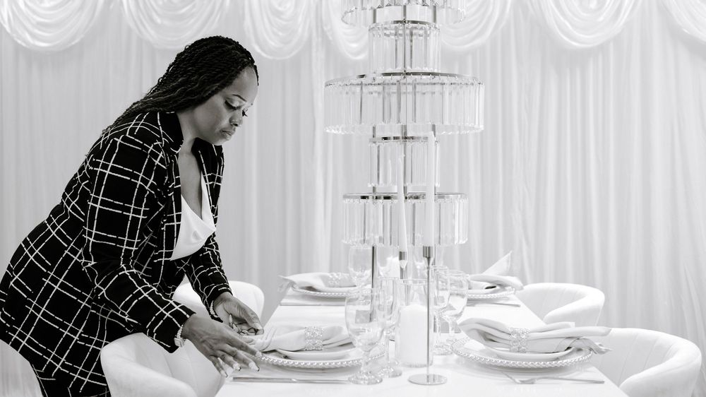 Woman in plaid blazer setting an elegant white table with crystal glassware and tall centerpiece.