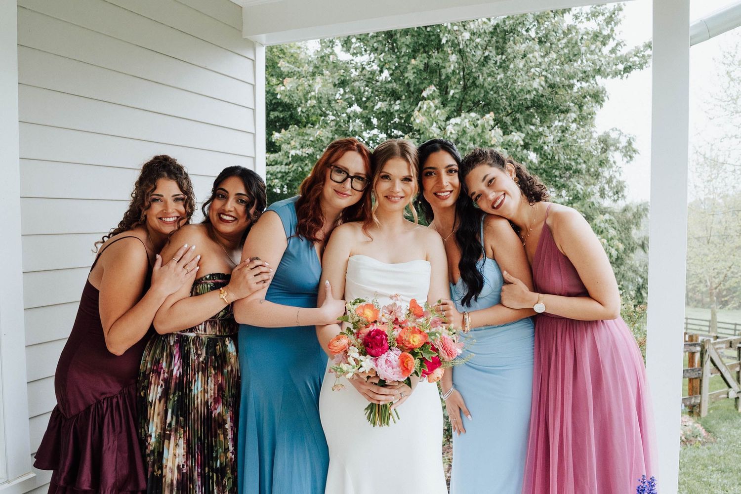 Bride in white gown holds colorful bouquet surrounded by smiling bridesmaids in blue and purple dresses on a porch.