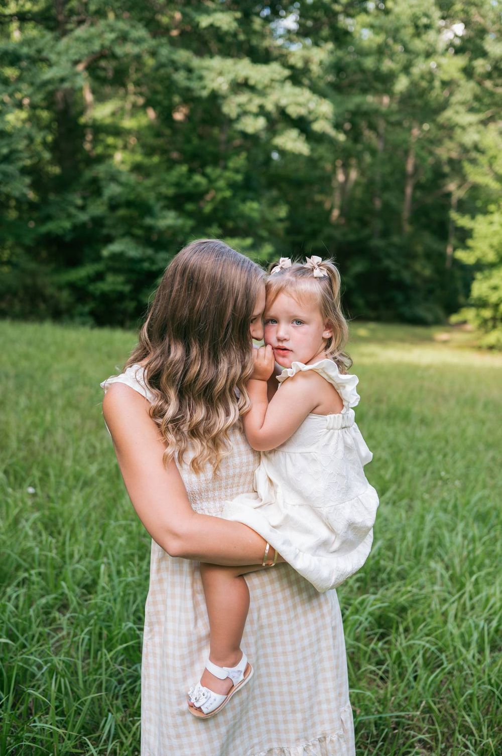 Mother kissing her toddler daughter on the cheek while holding her in a lush green meadow High Point NC